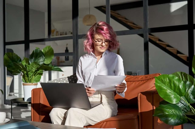 Young adult woman student with pink hair using laptop computer doing paperwork at home online on sofa in sunny cozy living room.