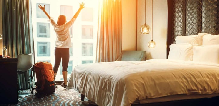 Woman in a hotel room, looking out the window, next to a bed